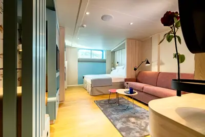 Innenansicht einer hellen Familienkabine mit Doppelbett, rosafarbenem Sofa, Holzboden und einem kleinen Fenster. 
