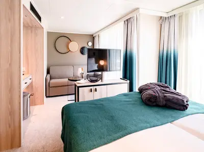 Blick in eine helle, moderne A-ROSA Suite der Kategorie F mit Bett, Sofa, TV, Badezimmereingang und Vorhängen in Türkis und Weiß.