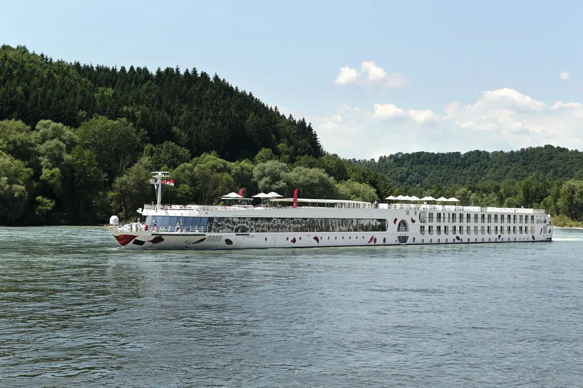 Das weiße Flussschiff A-ROSA SILVA mit roten Rosenmustern am Bug fährt auf dem Rhein, vorbei an grünen, bewaldeten Hügeln unter blauem Himmel.