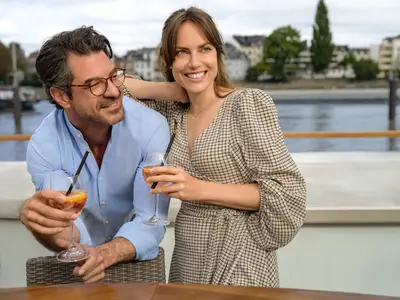 Ein lachendes Paar mit Aperitif-Gläsern auf dem Sonnendeck der A-ROSA SILVA. Im Hintergrund ist ein Fluss mit Bebauung zu sehen.