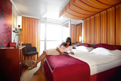Eine Frau liegt entspannt auf dem Bett einer warm eingerichteten A-ROSA STELLA Kabine mit französischem Balkon und liest ein Magazin.