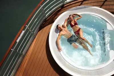 Ein Paar entspannt in einem runden Whirlpool auf einem Kreuzfahrtschiff. Im Hintergrund sind das Wasser und ein Geländer zu sehen.