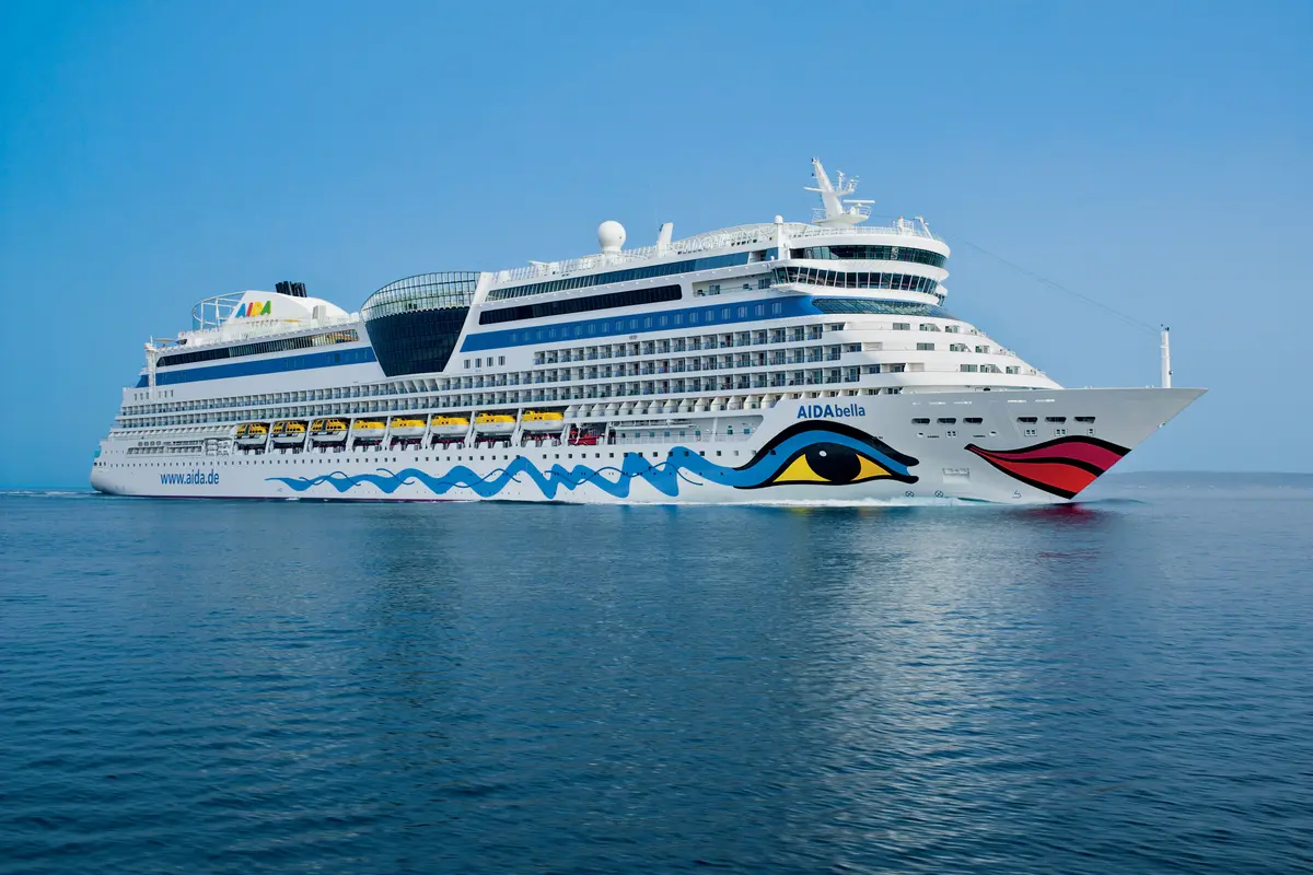 Die AIDAbella, ein großes Kreuzfahrtschiff, fährt auf dem offenen Meer unter blauem Himmel. Das markante Design mit Kussmund und Auge ziert den Bug der weißen Bordwand.