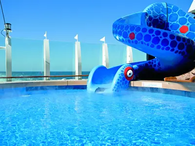 Das Ende einer blauen, monsterförmigen Wasserrutsche, die in einen Pool an Bord der AIDAbella mündet. Im Hintergrund sind das weite Meer und der blaue Himmel zu sehen, umrahmt von einer Glasbrüstung.