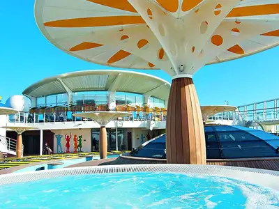 Sonniges Pooldeck der AIDAbella: Ein Whirlpool, pilzförmige Sonnenschirme und bunte Tanz-Silhouetten an der Wand. Im Hintergrund ein verglastes Restaurant. Blauer Himmel über der AIDAbella.