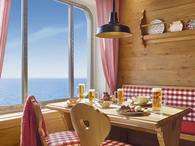 Gemütliches Restaurant an Bord der AIDAbella im Almhütten-Stil. Holztisch mit Bier, Brezeln und Speisen vor großem Fenster zum Meer. Rote karierte Vorhänge und Sitzbank.