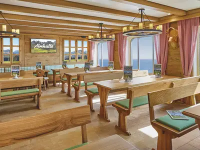 Innenansicht der rustikalen Almhütte an Bord der AIDAbella. Helle Holzbänke, grüne Kissen, Holztische. Links Berge, rechts Meer. Hirschkopf an der Wand.