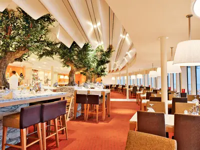 Das Bella Vista Restaurant an Bord der AIDAbella. Helle Einrichtung mit Olivenbäumen, Tischen, Buffet und Fenstern.