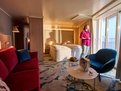In einer modernen Suite an Bord der AIDAbella blickt eine Frau durch die Balkontür. Die Kabine ist ausgestattet mit Doppelbett, rotem Sofa, blauem Sessel und einem Tisch mit Sekt und Früchten.