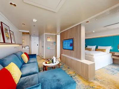 Geräumige Suite an Bord der AIDAbella mit einem blauen Ecksofa im Vordergrund und einem Doppelbett im Hintergrund. Ein heller Holztrennbereich mit Fernseher und ein Schreibtisch mit rotem Stuhl sind ebenfalls sichtbar. Die Einrichtung ist modern und hell.