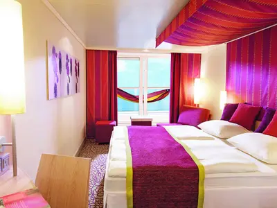 Geräumige Balkonkabine an Bord der AIDAblu. Ein bequemes Doppelbett, farbenfrohe Kissen und ein Balkon mit Hängematte sind zu sehen. Die Einrichtung ist modern und in Lila- und Rottönen gehalten.