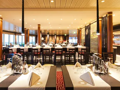 Blick in das geräumige East Restaurant auf der AIDAblu. Zahlreiche Tische sind weiss gedeckt mit Besteck, Gläsern und Servietten. Im Hintergrund große Fenster und Holzelemente.