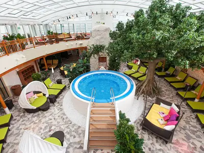 Vogelperspektive auf den großzügigen Body & Soul Spa der AIDAblu mit zentralem Whirlpool, vielen Liegen, grünen Pflanzen und einem hellen, überdachten Innenbereich.