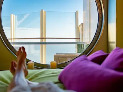 Eine Person entspannt auf einem Bett an Bord der AIDAcosma und blickt durch ein rundes Fenster auf das Meer und die Reling. Kerzen und violette Kissen schaffen eine gemuetliche Atmosphaere.