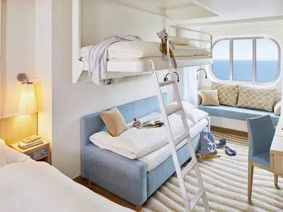 Geräumige Meerblickkabine an Bord der AIDAcosma mit weißem Bett, ausziehbarem Sofa, Hochbett mit Leiter und großem Fenster zum Meer. Ein Schreibtisch steht rechts.