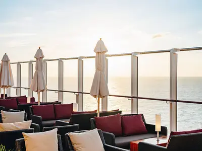 Blick auf die Aussenlounge der AIDAcosma mit gemuetlichen Sofas und Sonnenschirmen. Die Sonne scheint goldgelb ueber dem weiten Meer im Hintergrund.