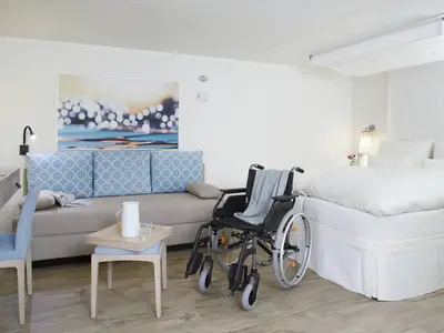 Eine helle, barrierefreie Verandakabine Komfort an Bord der AIDAcosma mit einem Doppelbett, einem Sofa mit Kissen, einem Schreibtisch und einem Rollstuhl auf Holzboden.