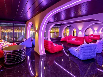 Blick in die moderne Teen Lounge Wave Club an Bord der AIDAcosma bei Nacht. Der Raum ist in Lila- und Rottönen gehalten, mit bequemen, farbigen Sitzsäcken und Sofas. Neonlichtstreifen beleuchten die Architektur. Durch große Fenster ist eine nächtliche Stadt-Skyline mit reflektierenden Lichtern im Wasser zu sehen. Der Boden glänzt schwarz und spiegelt die Lichter wider.