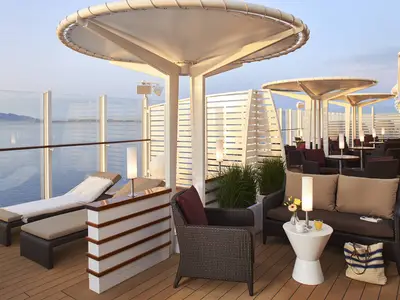 Aussen-Lounge an Bord der AIDAcosma. Holzdeck mit bequemen Rattan-Sitzmöbeln und Sonnenliegen. Glasbrüstung bietet weiten Meerblick auf ferne Berge. Schattensegel und dezente Lampen schaffen entspannte Abendstimmung.