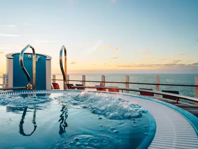 Ein sprudelnder Whirlpool auf dem Oberdeck der AIDAcosma bei Sonnenuntergang. Im Hintergrund erstreckt sich das weite Meer unter einem leicht bewölkten Himmel.
