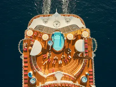 Vogelperspektive auf das Pooldeck der AIDAcosma. Ein großer Swimmingpool, zwei Whirlpools und viele rote Liegestühle auf hellem Holzdeck. Gelb-weiße Sonnenschirme und dunkles Meer sind sichtbar.