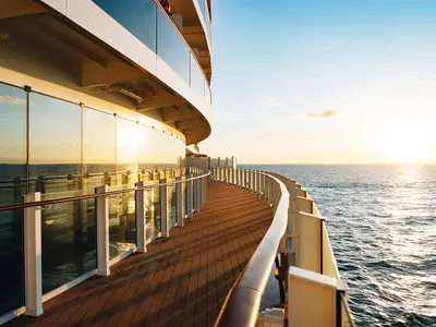 Die lange Promenade der AIDAcosma bei Sonnenuntergang. Goldgelbes Licht spiegelt sich auf dem Holzdeck, den Glasfassaden und dem weiten, glitzernden Meer auf der rechten Seite.