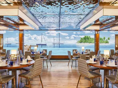 Das moderne Restaurant an Bord der AIDAcosma zeigt festlich gedeckte Tische. Ein riesiger Bildschirm simuliert einen Meerblick auf tropische Inseln mit Palmen und Segelyachten unter blauem Himmel.