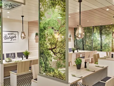 Modernes "Best Burger @SEA" Restaurant an Bord der AIDAcosma. Helle Holztische und Bänke, gedeckt. Grüne Baum- und Blättermotive auf Glaswänden. Pendelleuchten, Holzdecke. Keine Personen.