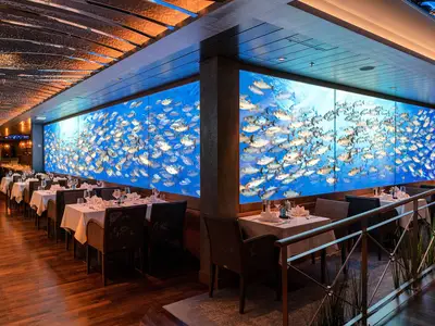 Blick in das Fischrestaurant Ocean's an Bord der AIDAcosma. Gedeckte Tische reihen sich vor einer riesigen, blau leuchtenden Digitalwand, die einen Fischschwarm zeigt.