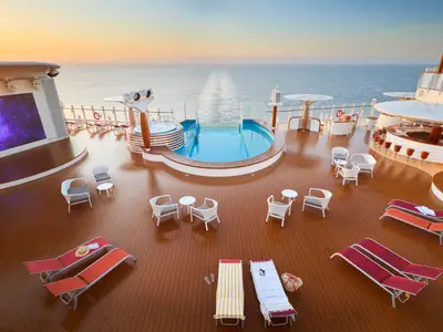 Weitwinkelaufnahme des sonnigen Holzdecks der AIDAcosma bei Sonnenaufgang mit ovalem Pool, Whirlpool, leeren Liegestühlen, Tischen und einer Bar. Ein großer Bildschirm zeigt Galaxien.