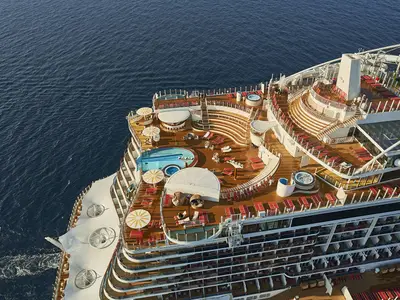 Luftaufnahme des weitläufigen Sonnendecks der AIDAcosma. Mehrere Pools, Whirlpools und rote Liegestühle auf Holzdecks sind zu sehen. Passagiere entspannen auf dem modernen Deck.