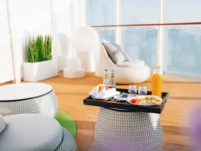 Blick auf ein modernes Outdoor-Deck an Bord der AIDAcosma. Auf einem Beistelltisch stehen ein Obstteller, Brot, Wasserflaschen und Orangensaft bereit. Bequeme Lounge-Sessel laden zum Entspannen ein, mit weitem Meerblick durch grosse Fenster.