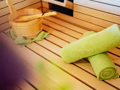 Nahaufnahme eines Spa-Wellness-Bereichs an Bord der AIDAcosma. Zu sehen sind helle Holzbänke, ein hölzerner Saunakübel mit Kelle und zwei gerollte grüne Handtücher, die auf den Bänken liegen.