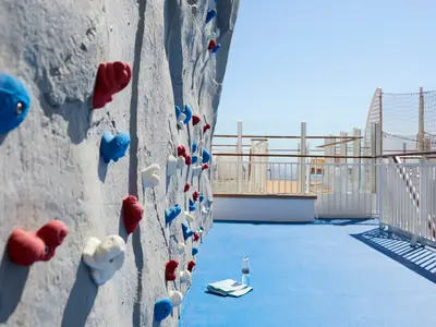 Kletterwand an Bord der AIDAcosma mit roten, blauen, weißen Griffen. Eine Wasserflasche und Handtuch liegen auf dem blauen Deck. Im Hintergrund sind Sportbereiche mit Netzen und das Meer unter klarem Himmel sichtbar.