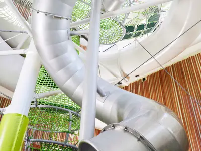 Blick auf eine moderne, helle Wasserrutsche im mehrstufigen Fun Park an Bord der AIDAcosma. Gitterstrukturen und grüne Netze sichern den Bereich. Im Hintergrund eine holzverkleidete Wand. Keine Personen sichtbar.
