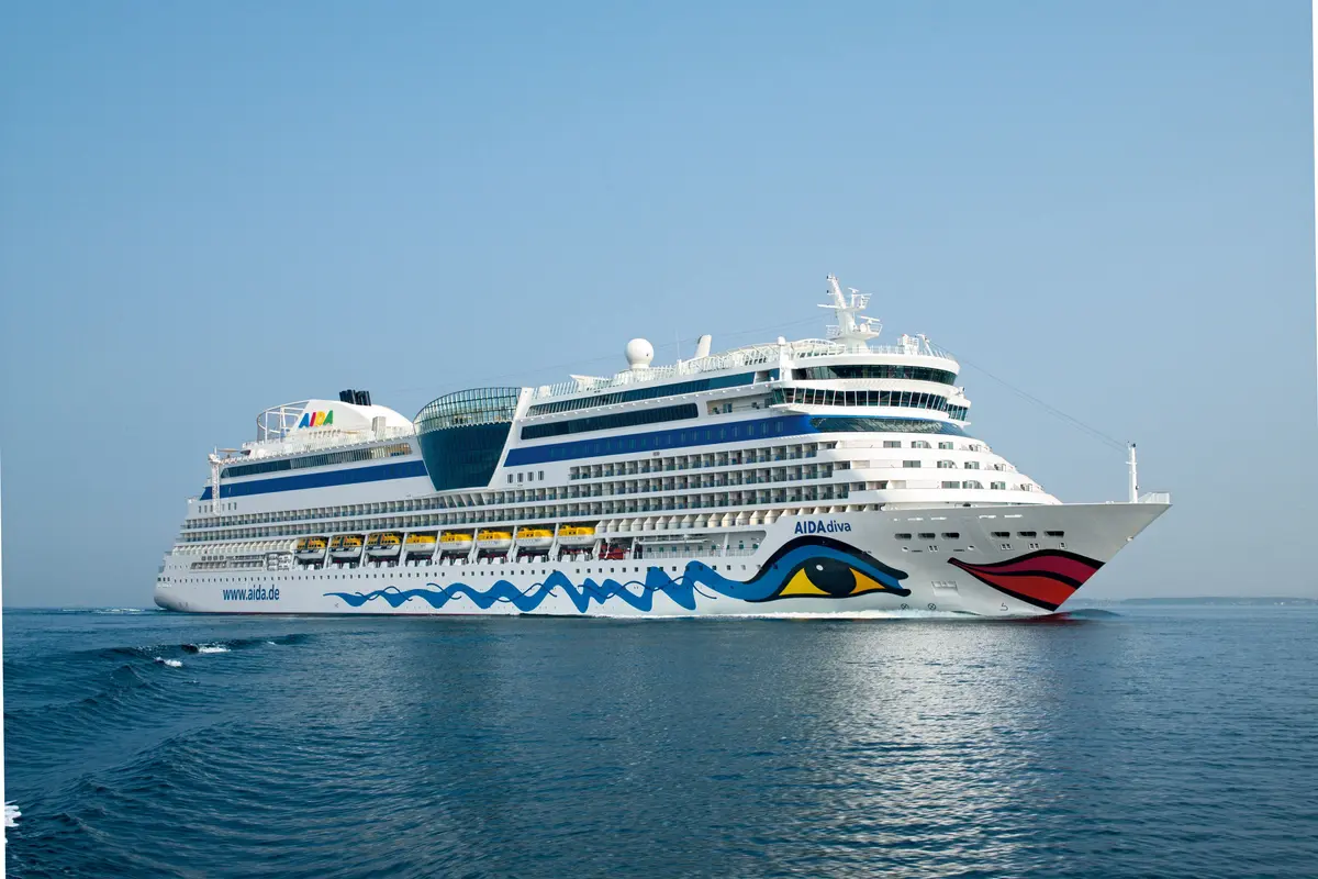 Die AIDAdiva auf dem offenen Meer. Das weisse Kreuzfahrtschiff mit Kussmund-Logo und blauen Wellenlinien am Rumpf gleitet friedlich unter blauem Himmel.