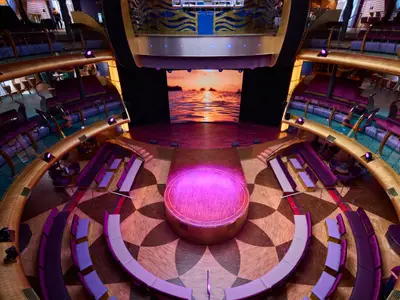Ansicht des AIDAdiva Theatriums. Bühne mit Sonnenuntergang auf LED-Wand. Magenta beleuchtete Rundbühne. Lila Sitzbereiche auf Ebenen.