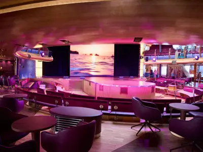 Weitwinkelansicht des Theatriums an Bord der AIDAdiva. Zu sehen sind eine Bühne mit grosser LED-Wand, davor runde Tische und Sessel in Lila. Die Raumbeleuchtung ist pinkfarben.