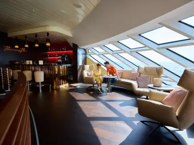 Die Panorama Bar auf der AIDAdiva mit stilvollem Barbereich und gemütlicher Lounge. Zwei Gäste unterhalten sich bei Tageslicht vor großen Fenstern mit Meerblick.