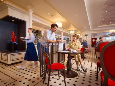 Ein gemütliches Café an Bord der AIDAdiva mit schwarz-weißem Fliesenboden und roten Stühlen. Ein Paar unterhält sich, während Crew Speisen serviert. Eine rote Skulptur ist sichtbar.