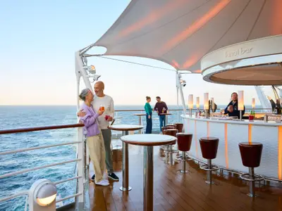 Ein älteres Paar trinkt Cocktails und umarmt sich an der Lanai Bar der AIDAdiva auf dem Außendeck bei Abendstimmung. Im Hintergrund ein Barkeeper und weitere Gäste.