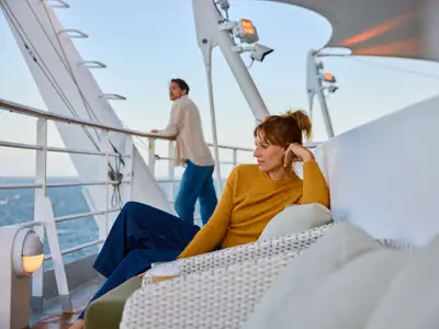 Eine Frau entspannt auf einer Liege an Deck der AIDAdiva, mit einem Getränk und Blick auf das weite Meer. Im Hintergrund lehnt ein Mann an der Reling und genießt die Aussicht.