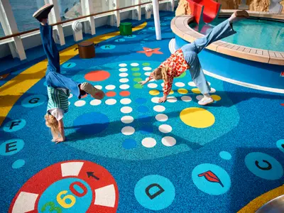 Zwei Kinder spielen auf dem farbenfrohen Außendeck der AIDAdiva, das als Spielbereich dient. Eines macht einen Handstand, das andere beugt sich nach vorne. Im Hintergrund ist ein Pool zu sehen.