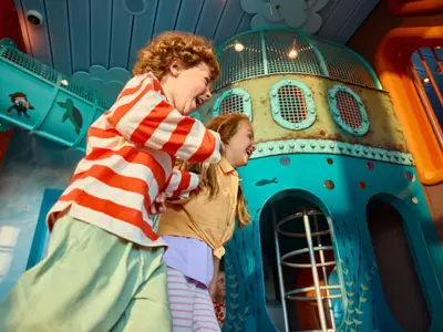 Zwei lachende Kinder spielen ausgelassen im fantasievollen Kids Club an Bord der AIDAdiva. Spielstrukturen im Unterwasser-Look wie ein U-Boot und eine Rutsche sind sichtbar.