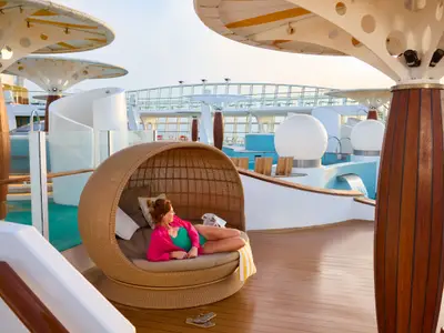 Eine Frau entspannt in einer Korbliege auf dem sonnigen Pooldeck der AIDAdiva. Im Hintergrund Pools mit Wasserspielen und moderne Überdachungen.