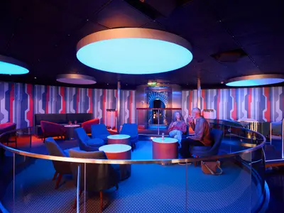 Zwei Gäste entspannen bei Drinks in der modern beleuchteten Bar-Lounge Oceans Club an Bord der AIDAdiva. Die Inneneinrichtung ist futuristisch mit blauen und roten Akzenten.