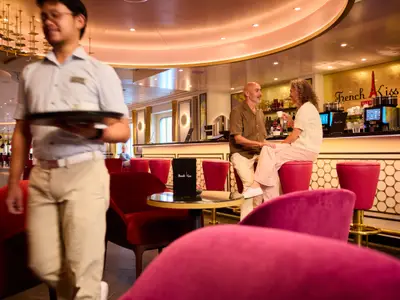 Ein Mann und eine Frau sitzen an der eleganten "French Kiss" Bar an Bord der AIDAdiva und trinken Kaffee. Ein Crew-Mitglied geht im Vordergrund. Rote Sessel und goldene Akzente prägen den Raum.