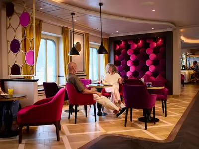 Blick in die elegante French Kiss Bar & Lounge der AIDAdiva mit Gästen an Tischen und einer Bar im Hintergrund. Das Interieur ist geprägt von roten Samtsesseln und goldfarbenen Vorhängen.
