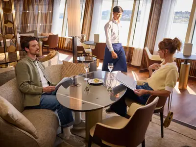 Ein Paar entspannt in einer stilvollen Lounge der AIDAdiva, liest Speisekarten und genießt den Meerblick durch große Fenster. Ein Kellner geht im Hintergrund vorbei.
