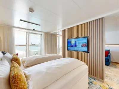 Innenansicht einer geräumigen Suite mit zwei Betten, Fernseher und Balkonzugang an Bord der AIDAdiva. Ein offener Wohnbereich ist rechts sichtbar.